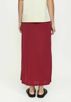 Isik, kes kannab kreemjat lühikese varrukaga t-särki ja pikka punast seelikut külje lõhikute ja mustade libisevate sandaalidega, seisab seljaga vaataja poole.