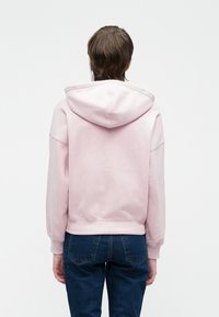 Person facing away wearing a light pink hoodie and dark blue jeans against a plain background.