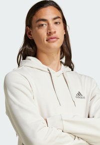 Vaaleanbeige huppari, jossa on pehmeä rakenne, nyöri hatussa ja valkoinen adidas-logo vasemmassa rinnassa. Pitkät hihat ja rento leikkaus.