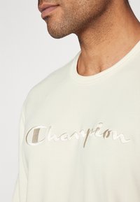 Gros plan d'une personne portant un t-shirt blanc à col rond avec le mot « Champion » brodé sur la poitrine.