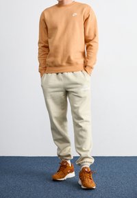 Person som bär en brun Nike sweatshirt, beige Nike sportbyxor och orange Nike sneakers som står på en blå matta.
