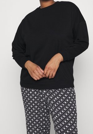 Person trägt ein schlichtes schwarzes Langarmshirt und schwarz-weiß gemusterte Hosen mit geometrischem Design, steht mit vor dem Körper verschränkten Händen.
