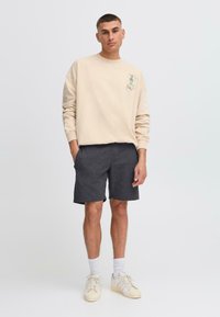 Sweatshirt beige avec un graphique sur la poitrine gauche, associé à un short gris foncé. Le mannequin porte des chaussettes blanches et des baskets claires, se tenant devant un fond blanc.