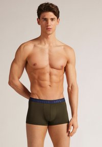 Olivgröna boxerkalsonger med en slät yta och marinblå elastisk midjeband som pryder varumärkesnamnet. Fitted design med en formad ficka.