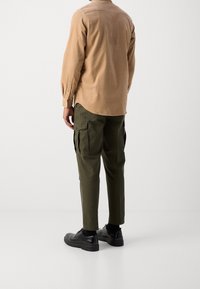 Hellbraun langärmliges Hemd mit Knopfmanschetten, kombiniert mit olivgrünen Cargo-Hosen mit seitlichen Taschen und schwarzen Schnürschuhen.