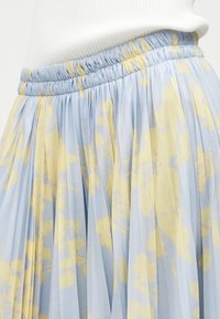 Φούστα σε γαλάζιο χρώμα με πιέτες και ανοιχτές κίτρινες ανθισμένες λεπτομέρειες, με ελαστική μέση, φορεμένη με λευκή ριμπ μπλούζα.