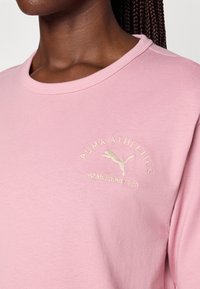 Hellsrose Baumwoll-T-Shirt mit rundem Halsausschnitt, kurzen Ärmeln und einem goldgestickten Logo mit dem Text "PUMA ATHLETICS gegründet 1948."