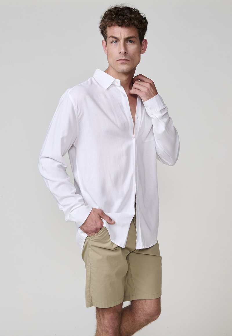 Cămașă albă cu mâneci lungi, cu nasturi, confecționată dintr-un material ușor, asociată cu pantaloni scurți khaki. Cămașa are un buzunar frontal și o croială lejeră.