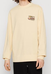 Sudadera color crema con mangas largas, cuello redondo y un gráfico en el pecho que presenta montañas y sol. Textura suave, ajuste holgado.