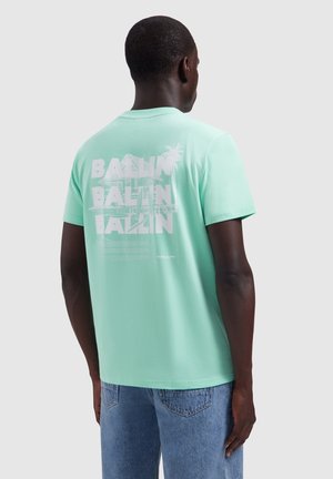 Man draagt een mintgroen T-shirt met witte tekst en een palmboomgrafiek op de achterkant, staand tegen een effen achtergrond.