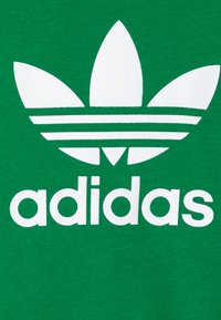 Zelená bavlněná tkanina s bílým logem Adidas trefoil, které má tři listy a tři vodorovné pruhy nad názvem značky v bílé.