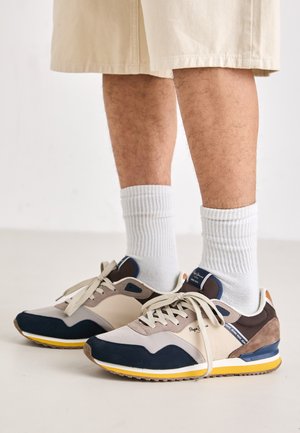 Sneakers con una tomaia in suede beige, blu e marrone, dotate di una suola gialla, lacci bianchi e calze bianche a coste.