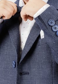 Niño ajustando un blazer de textura navy sobre una camisa blanca con lunares multicolores, enfocándose en las manos y los detalles de la ropa.