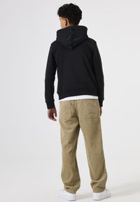 Sudadera de algodón negra con una gran capucha, combinada con pantalones de pana beige que tienen bolsillos traseros, y zapatillas blancas. Fondo neutro.