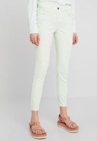 Vaqueros skinny de un ligero verde menta, hechos de denim, con cierre de botón en la parte frontal, cinco bolsillos y sutiles detalles de costura. Combinados con sandalias rosas.