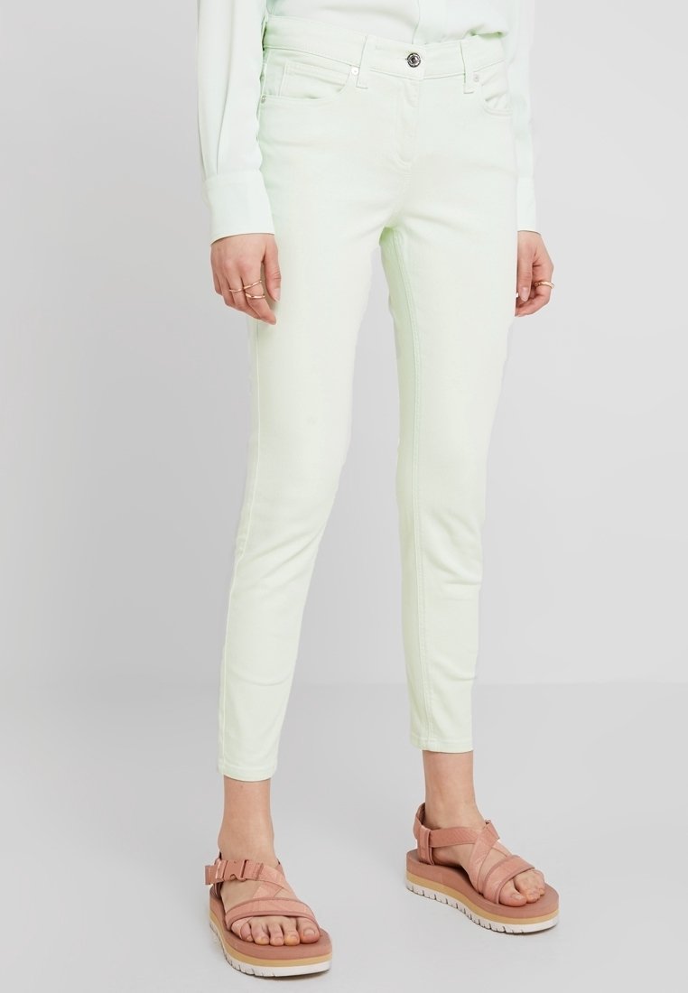 Vaqueros skinny de un ligero verde menta, hechos de denim, con cierre de botón en la parte frontal, cinco bolsillos y sutiles detalles de costura. Combinados con sandalias rosas.