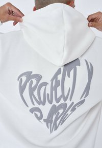 Sudadera blanca con un gráfico en gris que dice "PROJECT X" y un diseño de corazón en la parte posterior. La tela parece suave con una textura cepillada.