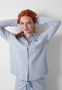 Blåt pyjamas sæt med et stribet mønster, der har en mønstret lomme med et Snoopy design. Skjorten har knaplukning foran og krave.