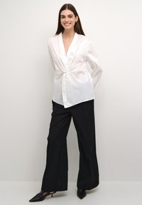 Camicia bianca avvolgente con scollo a V e dettaglio a fiocco, abbinata a pantaloni neri a vita alta e gamba larga, e décolleté neri a punta. Tessuto dalla texture liscia.