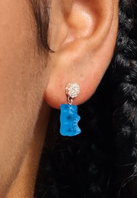 Ohr mit einem baumelnden Ohrring, der eine kleine Ansammlung klarer Edelsteine und einen durchscheinenden blauen Gummibärchen-Anhänger zeigt.