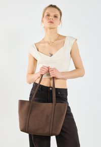 Borsa tote in pelle marrone con una superficie liscia, design strutturato e manici doppi. Modella tiene la borsa indossando una maglia chiara.