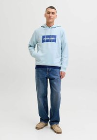 Jeune homme debout, une main dans la poche, portant un sweat à capuche bleu clair avec un logo, un jean bleu et des chaussures décontractées beiges sur fond blanc.