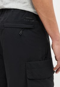 Άνδρας που φοράει μαύρα cargo σορτς με τσέπες με φερμουάρ και καπάκια, στέκεται με το χέρι χαλαρά στο πλάι.