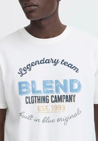 Balta medvilninė marškinėlių su spausdintu mėlynu ir juodu grafinio teksto įvaizdžiu, įskaitant "BLEND Clothing Company" ir "EST. 1993," su laisvu pjūviu.