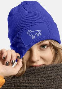 Blue knitted beanie with ribbed texture, featuring a rectangular patch with a white dog silhouette. Soft material and snug fit.