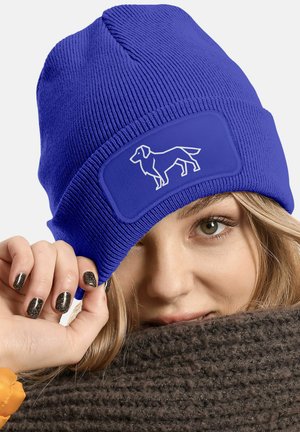 Blue knitted beanie with ribbed texture, featuring a rectangular patch with a white dog silhouette. Soft material and snug fit.