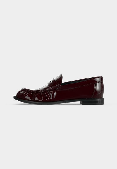 Mocassin en cuir bordeaux foncé brillant avec un petit talon et des détails froncés sur le bout, montré de profil sur un fond uni.