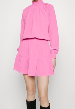 Žena v jasně růžových dlouhých šatech s volánkem u vysokého límce a vrstvenou sukní, doplněná černými kozačkami po kolena.