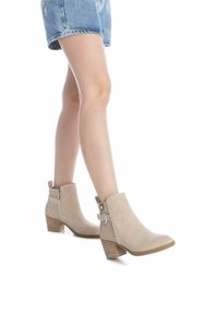 Botas de tobillo beige con un acabado texturizado, cremallera lateral, costuras decorativas y un tacón apilado. Llevadas con pantalones cortos de mezclilla.