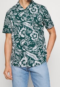 Vihreä lyhythihainen paita, jossa on valkoinen kukkakuosi. Paita on varustettu v-pääntiellä ja napituksella. Pehmeä kangas ja rento istuvuus.