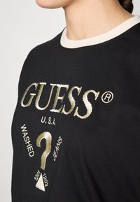 Svart bomullst-skjorte med en beige ribbet krage. Har gullbrodert tekst "GUESS®", "U.S.A." og "WASHED JEANS" med dekorative elementer.