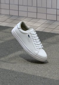 Hvit lav sneaker med snørebånd, svevende over grå asfalt nær flislagt vegg, viser side- og toppdetaljer med liten blå etikett.