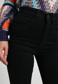 Zwarte high-waisted jeans hebben voorzakken en een gladde textuur. De stof is rekbaar, aansluitend en past perfect bij een veelkleurige gebreide trui.