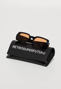 Gafas de sol retro en negro con lentes naranjas, exhibidas sobre un estuche suave negro que presenta el texto "RETROSUPERFUTURE" en blanco.
