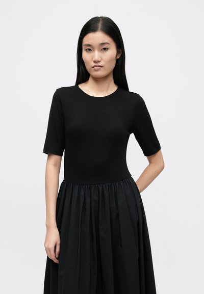 Čierne šaty s priliehavým živôtikom a krátkymi rukávmi, rozšírenou skladanou sukňou. Vyrobené z hladkej, ľahkej látky.