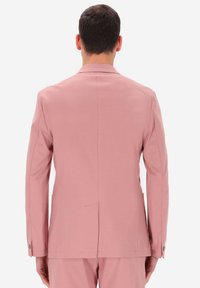 Uomo vestito con un completo rosa, visto di spalle, in piedi di fronte a uno sfondo bianco semplice.