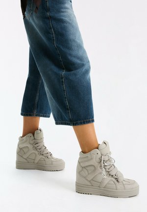 Person, die hellgraue High-Top-Sneakers und gekürzte blaue Jeanshosen trägt und auf einer weißen Oberfläche steht.