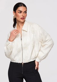 Femme aux cheveux foncés portant une veste blanche à motifs floraux texturés et à fermeture éclair, associée à un pantalon noir taille haute, accessoirisée avec de grandes boucles d'oreilles dorées.