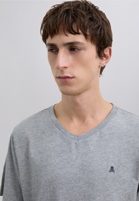 Graues V-Ausschnitt-T-Shirt mit kurzen Ärmeln, weicher Textur, mit einem kleinen dunkelblauen Totenkopf-Logo auf der linken Brust. Schlichtes Design und lässige Passform.