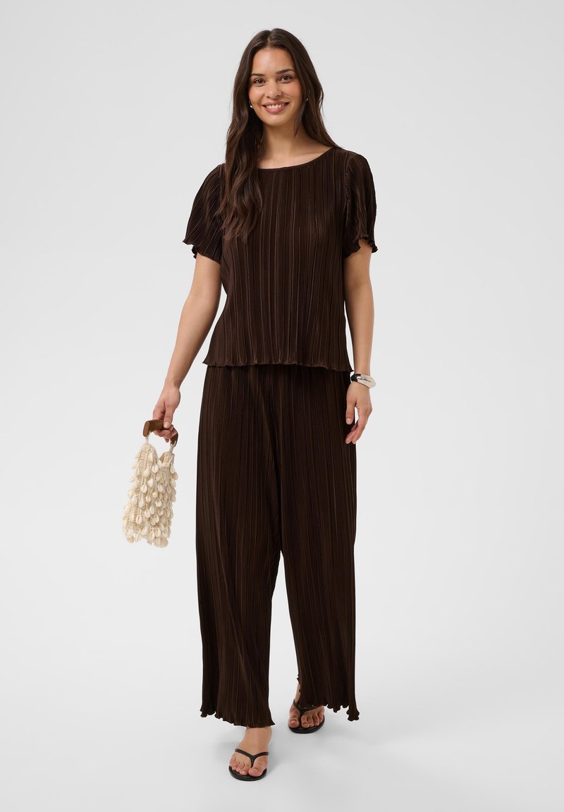 Femme souriante portant un haut plissé marron foncé assorti et un pantalon large, tenant un sac à main crème texturé et portant des tongs noires.