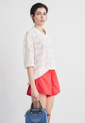 Jonge vrouw met kort donker haar, een wit shirt met patroon, rode korte broek, gouden oorring en een blauwe Adidas-handtas vasthoudend.