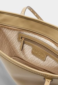 Bézs bőr bevásárlótáska belső cipzáras zsebbel és ismétlődő mintájú, texturált béléssel. Arany színű fém kiegészítőkkel díszítve.