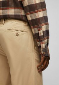 Braun- und beige-karierter Flanellhemd mit einem geknöpften Bündchen, kombiniert mit beigen Hosen, die eine Gesäßtasche mit Knopfdetail aufweisen.