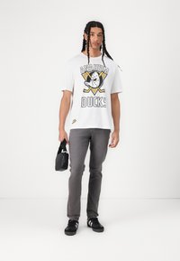 T-shirt grafica bianca con logo e elementi di design dei "Anaheim Ducks", abbinata a jeans grigi e sneaker nere. Il modello tiene una borsa nera.
