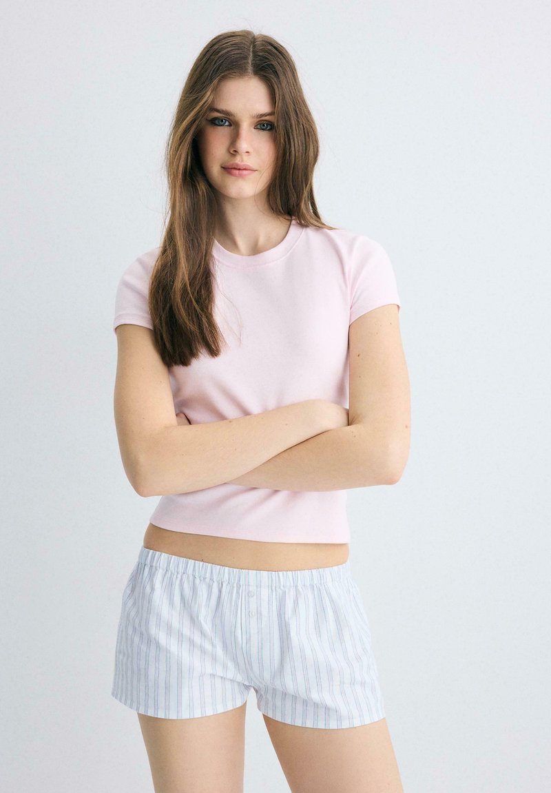 Jeune femme aux longs cheveux bruns portant un t-shirt rose clair et un short blanc à rayures, debout, les bras croisés, sur un fond gris uni.