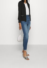 Černý blazer se stříbrným hardwarem, bílým tričkem, modrými džíny, stříbrnými lodičkami na podpatku a stříbrnou korálkovou kabelkou. Moderní casual outfit.